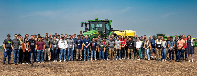 large group of students and professionals lined up in front of a tractor in a field.